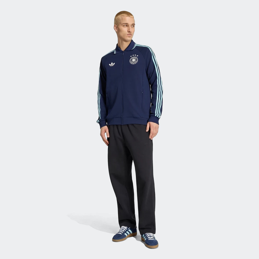 Male model wearing a navy blue Adidas Germany Away Originals track jacket with light blue stripes, black pants, and navy