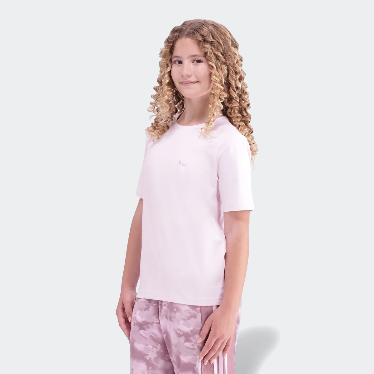 Girl with curly hair wearing a pink Adidas Premium Essentials Tee and pink camouflage pants, standing against a white