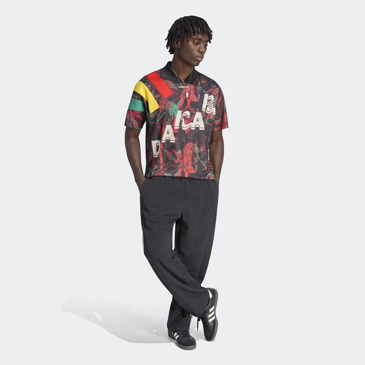 Black man wearing a black, red, green, and yellow Adidas Graphic Football Jersey, black pants, and black sneakers, full body