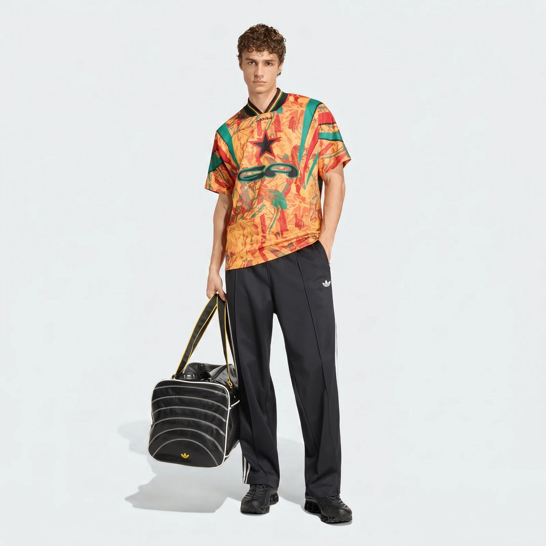 Full-body shot of a male model wearing a colorful graphic football jersey, black Adidas track pants, and holding a black