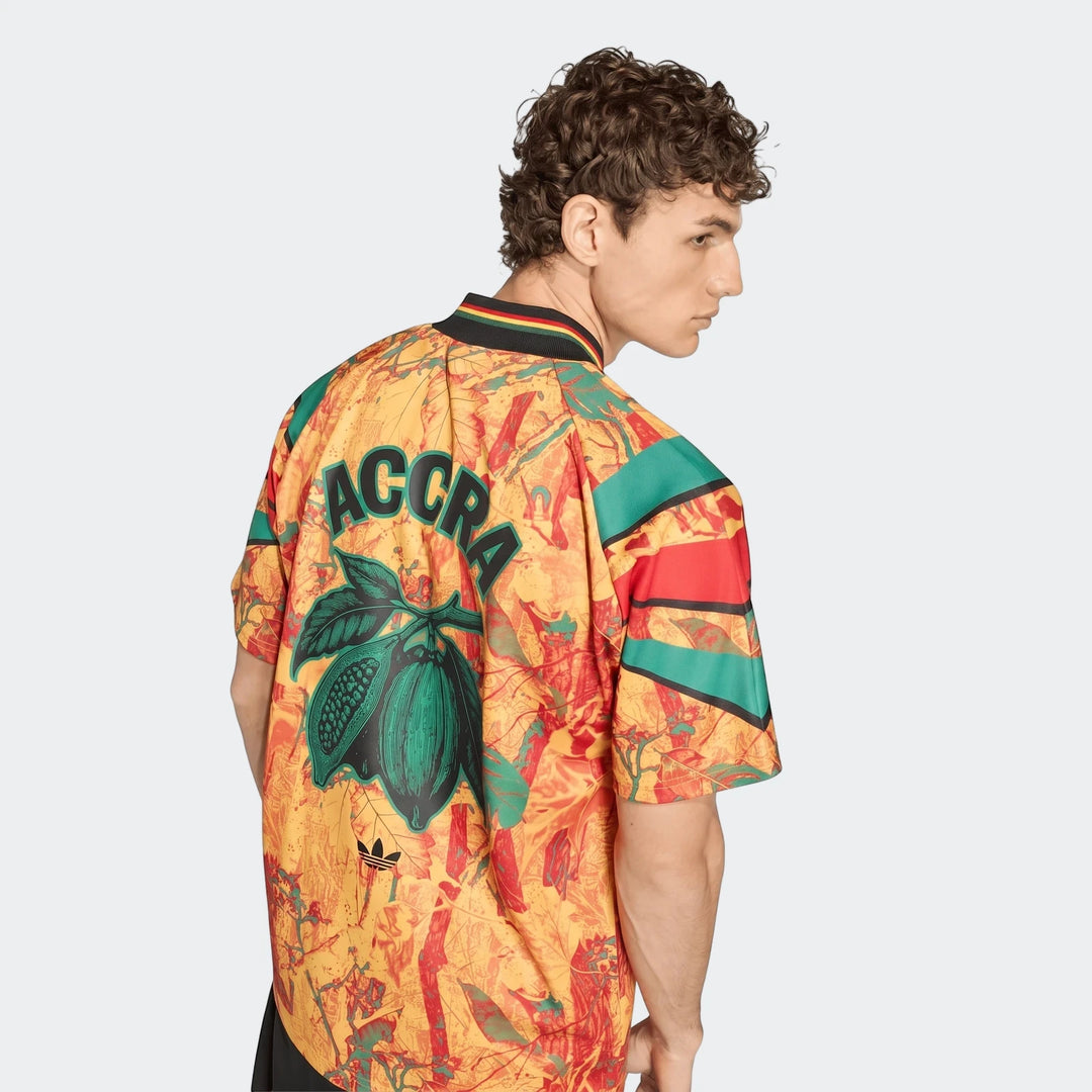 Male model wearing a yellow, red, and green graphic football jersey with 'Accra' text and a cocoa pod design on the back.