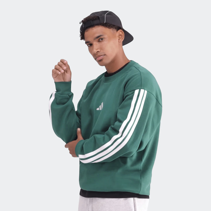 Male model in a green Adidas long-sleeve sweatshirt with white stripes and a black cap, posing from the waist up.