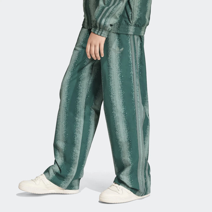 Green Adidas Sst Loose Track Pants with a unique pixelated stripe pattern, worn by a model in a studio shot.