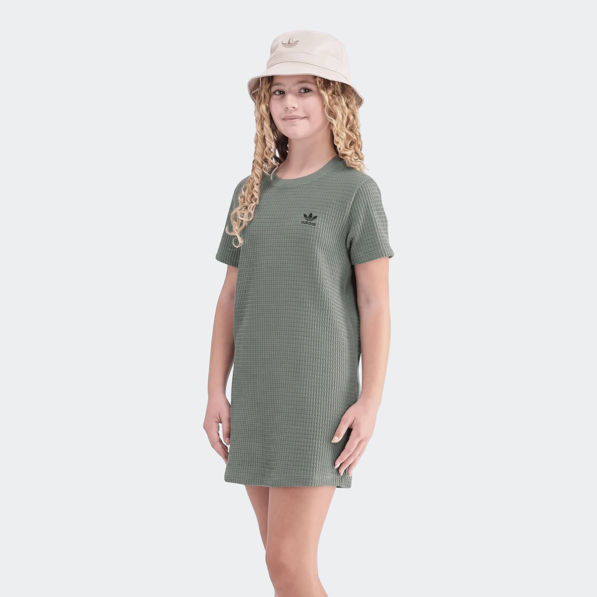 Young girl with curly blonde hair modeling a green Adidas waffle dress and a beige Adidas bucket hat, full body shot.