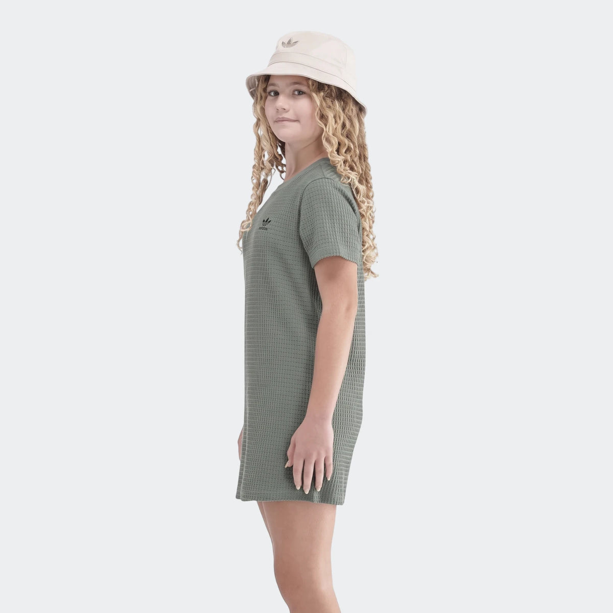 Young girl wearing a green Adidas waffle knit short-sleeve dress and a beige Adidas bucket hat, posing casually.