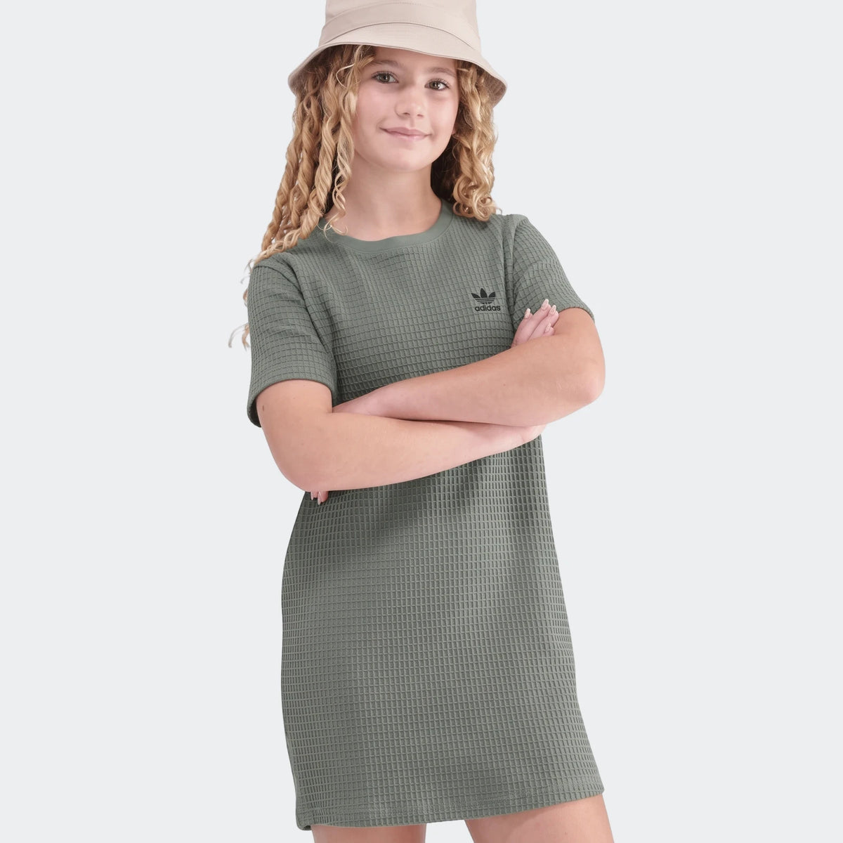 Young girl wearing a green short-sleeved waffle dress with an Adidas logo and a beige bucket hat, posing with arms crossed.