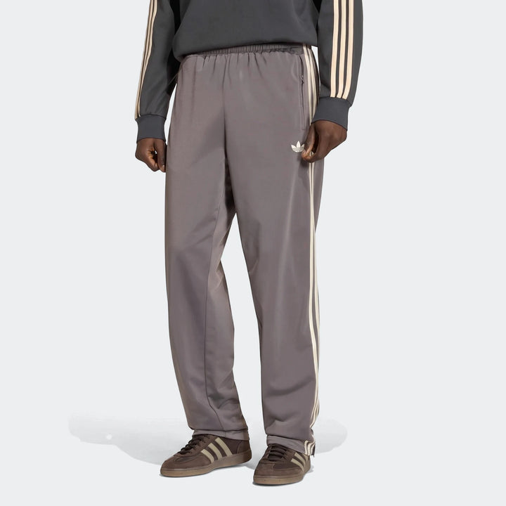Male model showcasing grey Adidas Firebird Track Pants with white stripes and logo, paired with brown sneakers, full length.