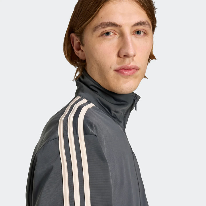 Close-up of a male model wearing a grey Adidas Firebird track top with white stripes on the sleeves.