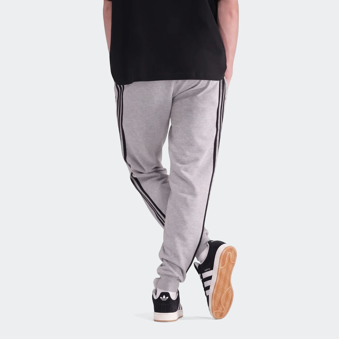 Grey men's 3-Stripes Pants, made of soft fabric, worn with black sneakers, viewed from the back with legs crossed.