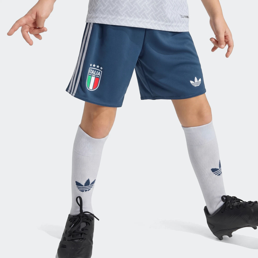 Young player in Italy 25/26 Away Mini Kit: blue shorts with team crest, white patterned jersey, white socks with Adidas logo.