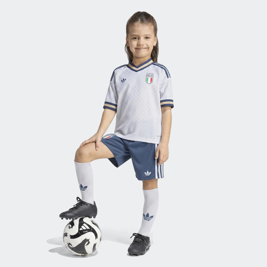 Young girl proudly wearing the Italy 25/26 Away Mini Kit, featuring a white jersey, blue shorts, and black cleats, with a