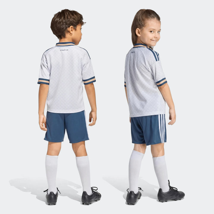 A young boy and girl wearing the Italy 25/26 away mini kit, featuring white shirts with blue shorts, viewed from the back.