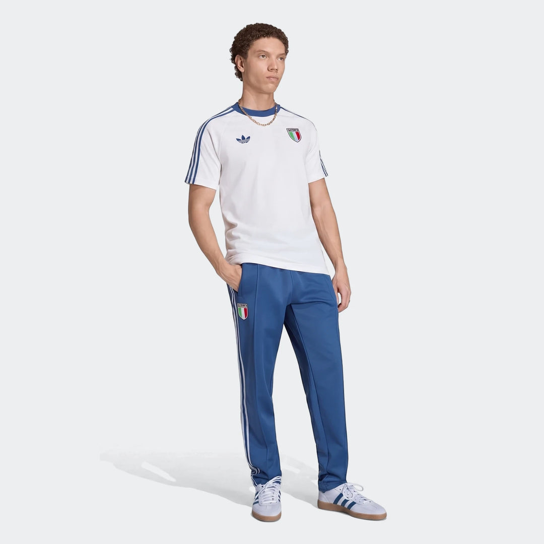 Male model wearing a white Adidas Italy Originals Graphic T-Shirt, blue track pants, and white sneakers posing in a studio.