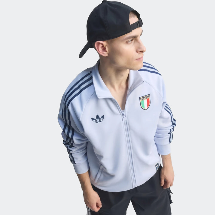 Waist-up shot of a male model wearing a light blue Adidas Italy Originals Track Top with dark blue stripes and a black cap.