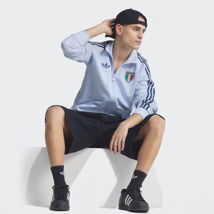 Young man modeling a light blue Adidas Italy Originals Track Top, black shorts, and black sneakers, seated in a studio.