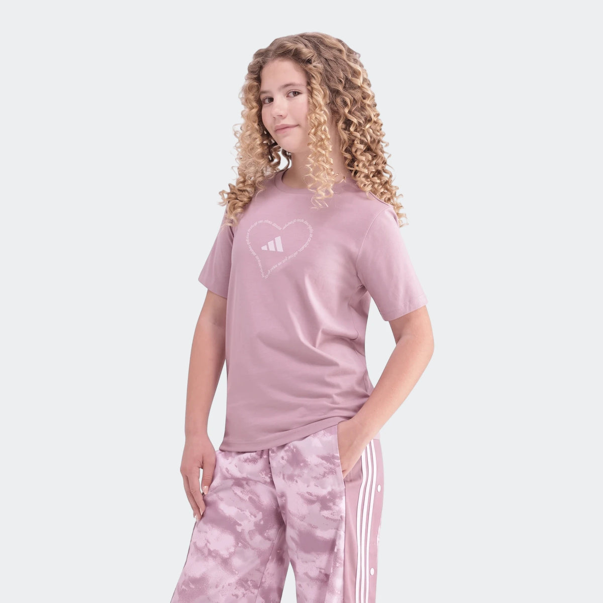 A young girl with curly hair models a light purple Adidas Jg Satin T-shirt with a heart logo and matching tie-dye sweatpants.