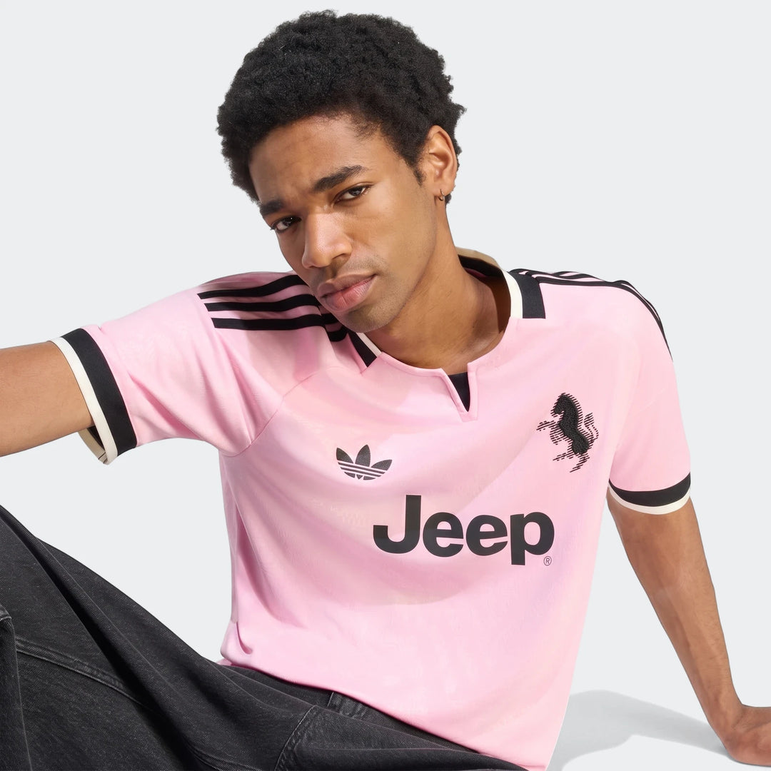 A male model wears a pink Juventus 26/27 Away Jersey with black stripes and logos, sitting and looking forward.