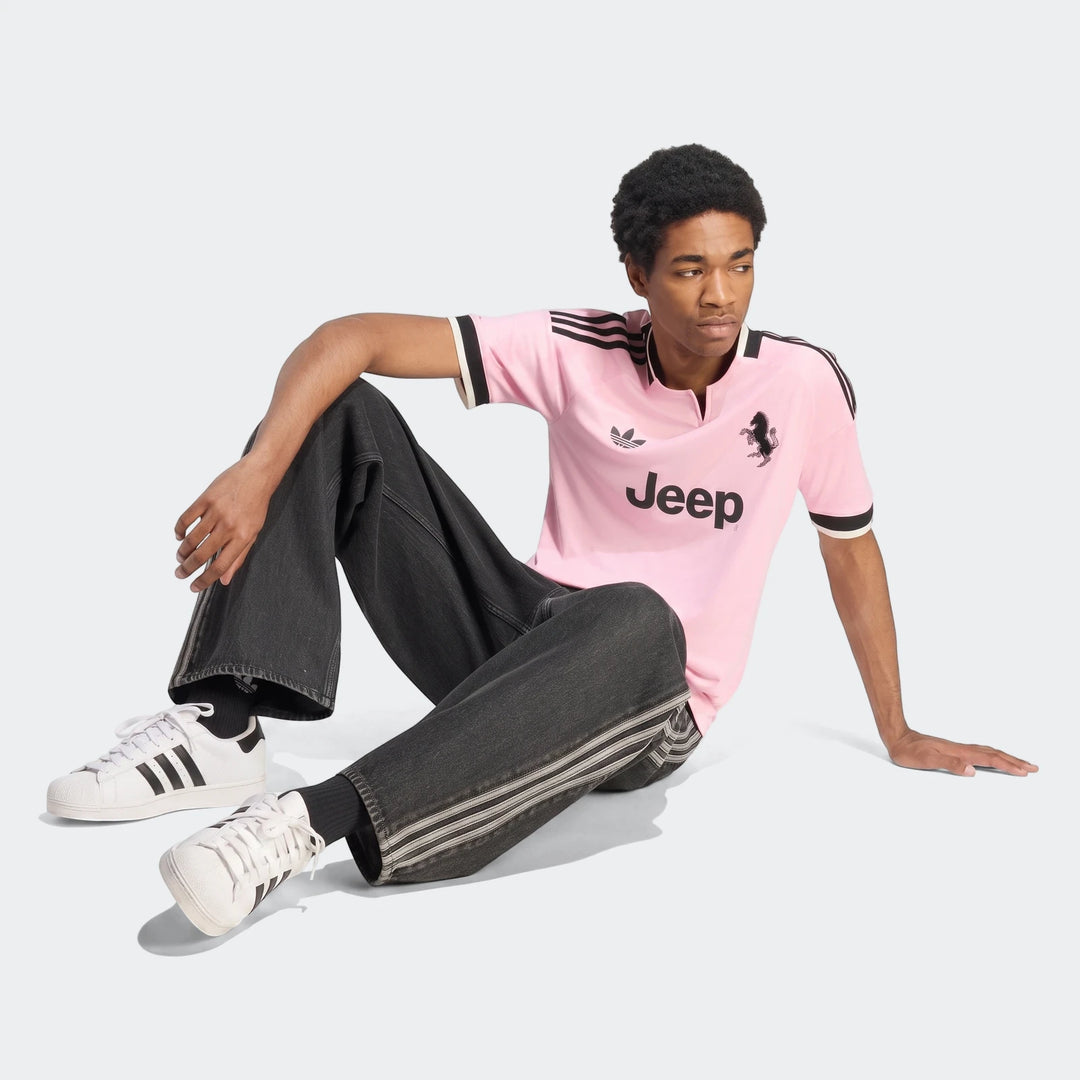 Male model wearing a pink Adidas Juventus 26/27 Away Jersey, dark grey jeans, and white Adidas Superstar sneakers, sitting
