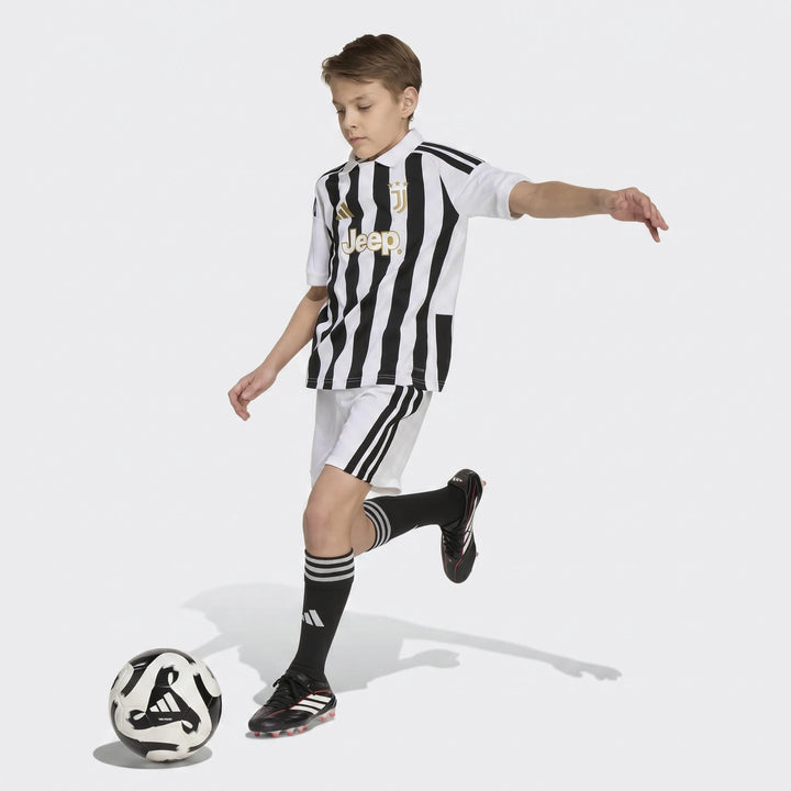 Young boy in black and white striped Juventus 26/27 Home Jersey, white shorts, and black socks, kicking a soccer ball.