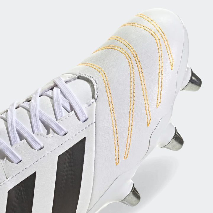 Close-up of white Kakari Elite SG rugby boot with black stripes and yellow stitching, showing laces and metallic studs.