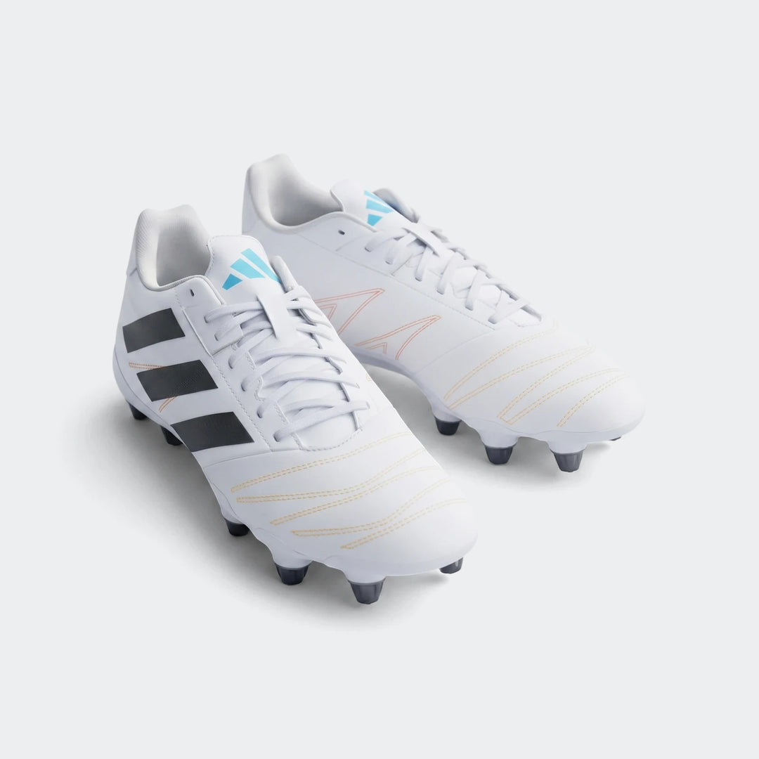 A pair of white Adidas Kakari Elite SG Rugby Boots with black stripes and blue logos, displayed on artificial green turf.