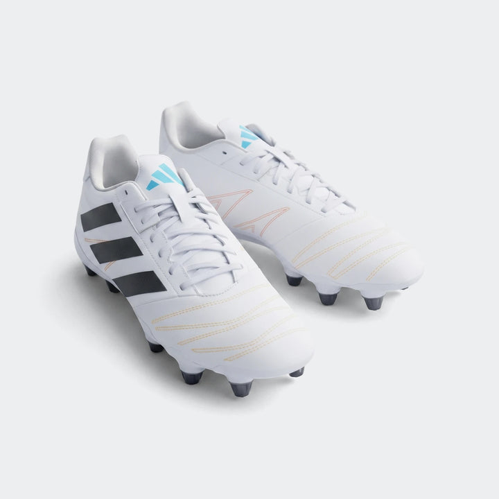 A pair of white Adidas Kakari Elite SG Rugby Boots with black stripes and blue logos, displayed on artificial green turf.