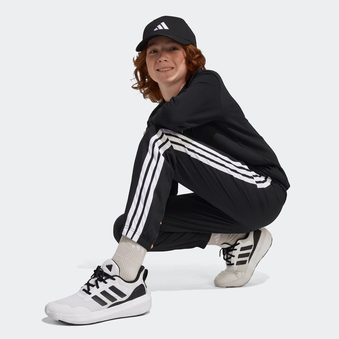 Black Adidas Essentials Climacool Tracksuit Bottoms for kids, featuring three white stripes, worn by a young person in a