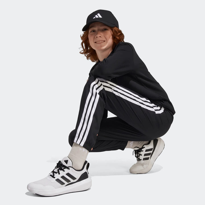 Black Adidas Essentials Climacool Tracksuit Bottoms for kids, featuring three white stripes, worn by a young person in a