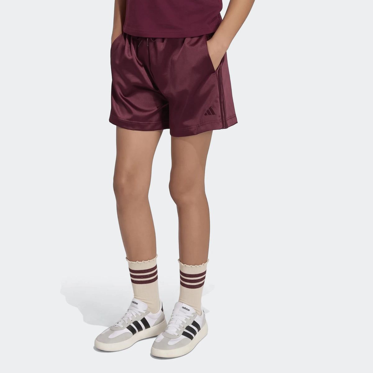 Kids burgundy Adidas satin shorts with white and grey sneakers and striped beige socks, modeled from the hips down.