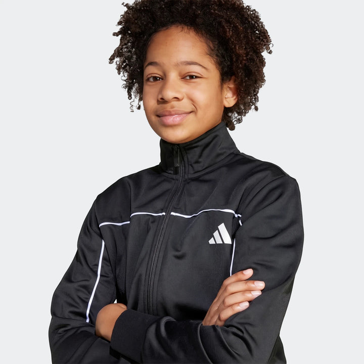 Young person wearing a black Kids Tricot Colour Pop Track Suit with white stripes and Adidas logo. Half-body shot of the