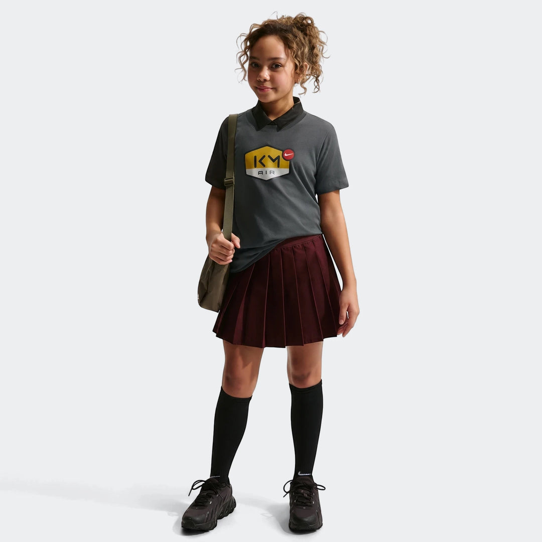 Young girl wearing a grey Nike Km K Nk Df Mbappe Tee, paired with a pleated burgundy skirt, black socks, and black sneakers.