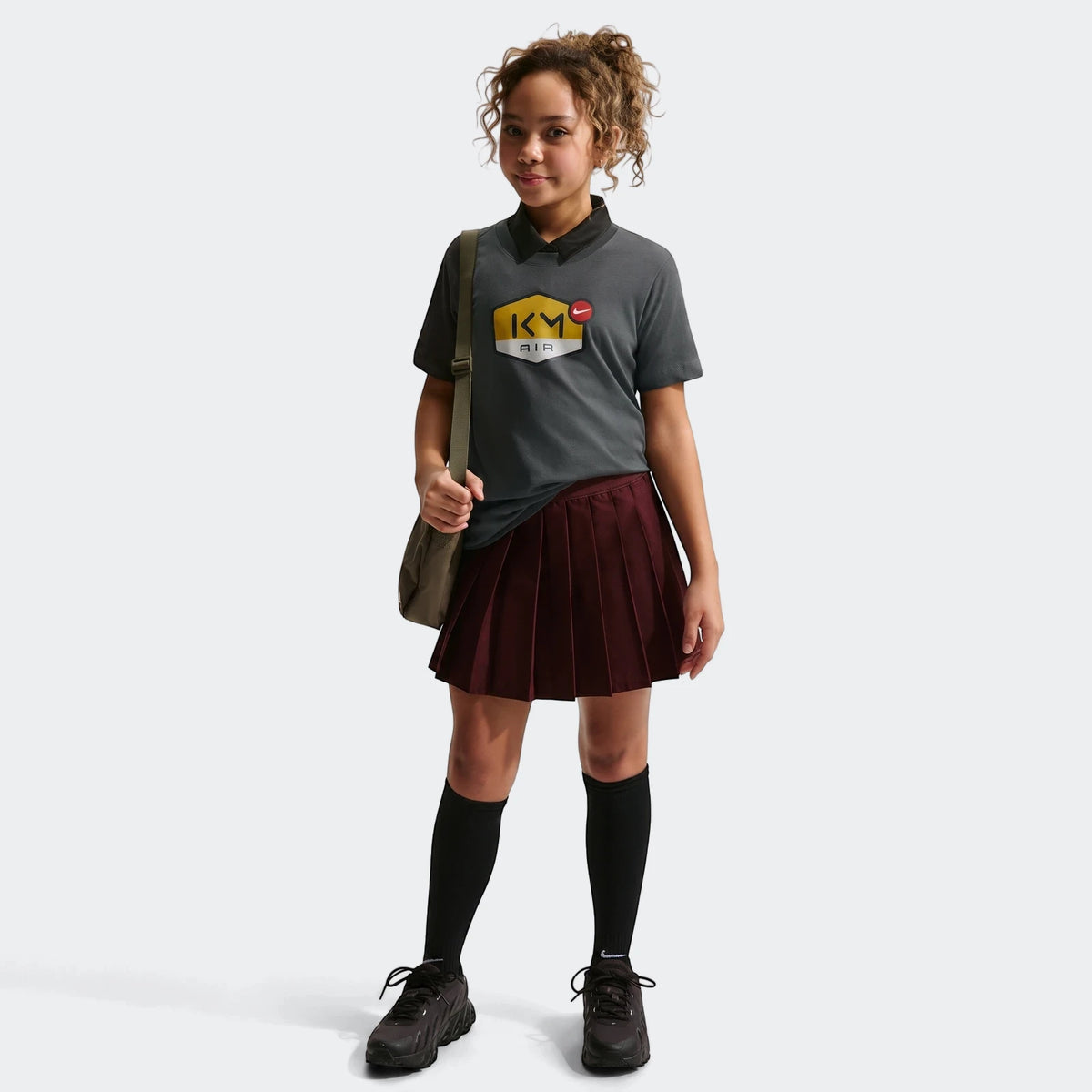 Young girl wearing a grey Nike Km K Nk Df Mbappe Tee, paired with a pleated burgundy skirt, black socks, and black sneakers.