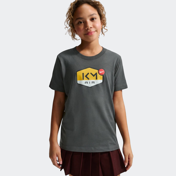 Young girl wearing a dark grey Km K Nk Df Mbappe Tee featuring a yellow and black graphic with a red Nike swoosh.