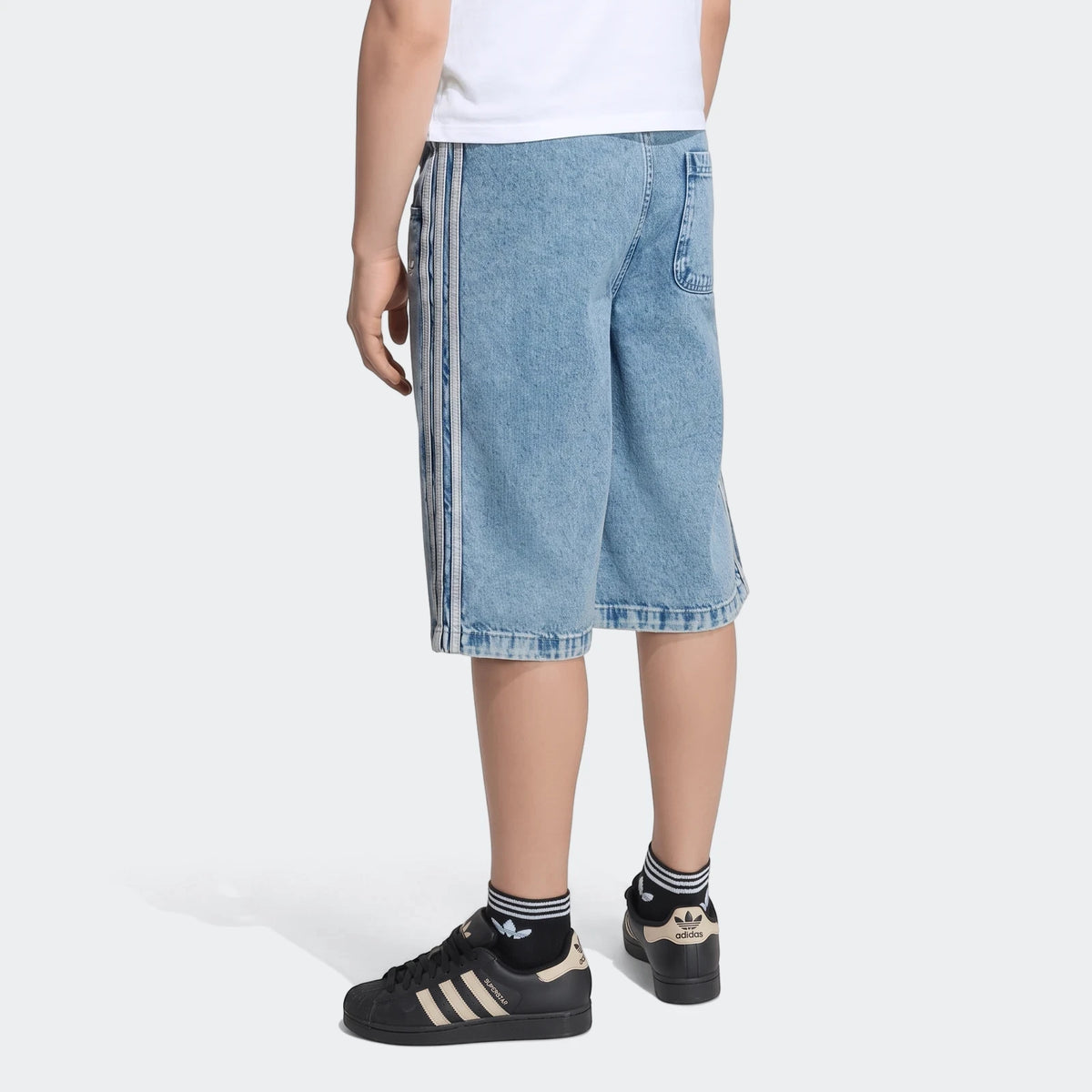 Rear view of a person wearing light blue loose denim jorts with white side stripes, black Adidas Superstar sneakers, and