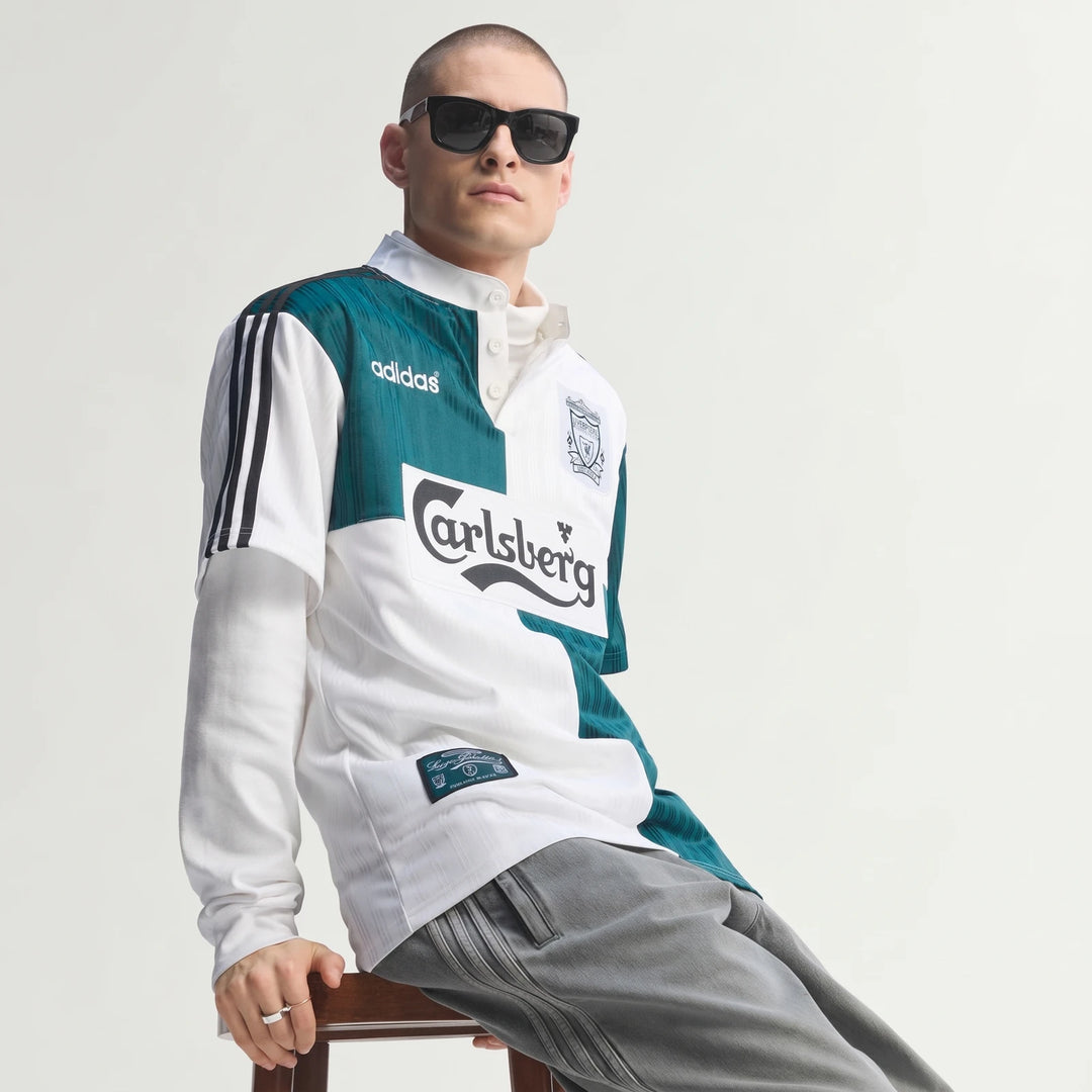 Male model wearing a white and teal Liverpool Fc 1995 Away Jersey with Adidas and Carlsberg logos, seated on a stool.