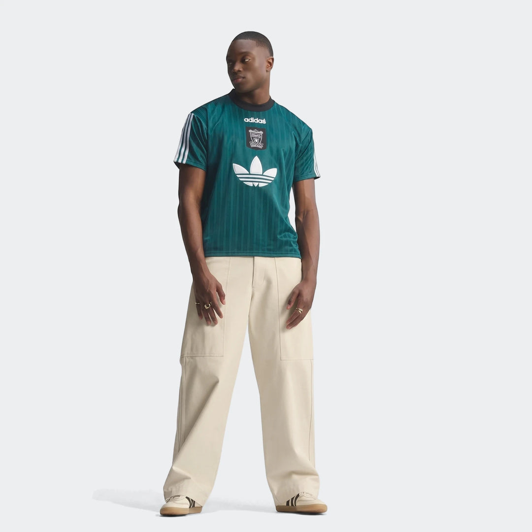 Male model wearing a dark teal green Liverpool Fc 1995 Poly T-Shirt, beige wide-leg pants, and sneakers, standing front view.