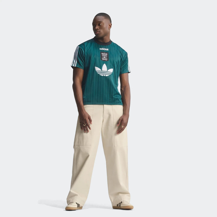 Male model wearing a dark teal green Liverpool Fc 1995 Poly T-Shirt, beige wide-leg pants, and sneakers, standing front view.
