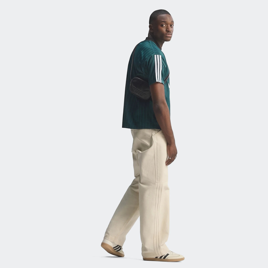 Man wearing a dark green Liverpool FC 1995 Poly T-Shirt with white stripes, light pants, and white sneakers, holding a black