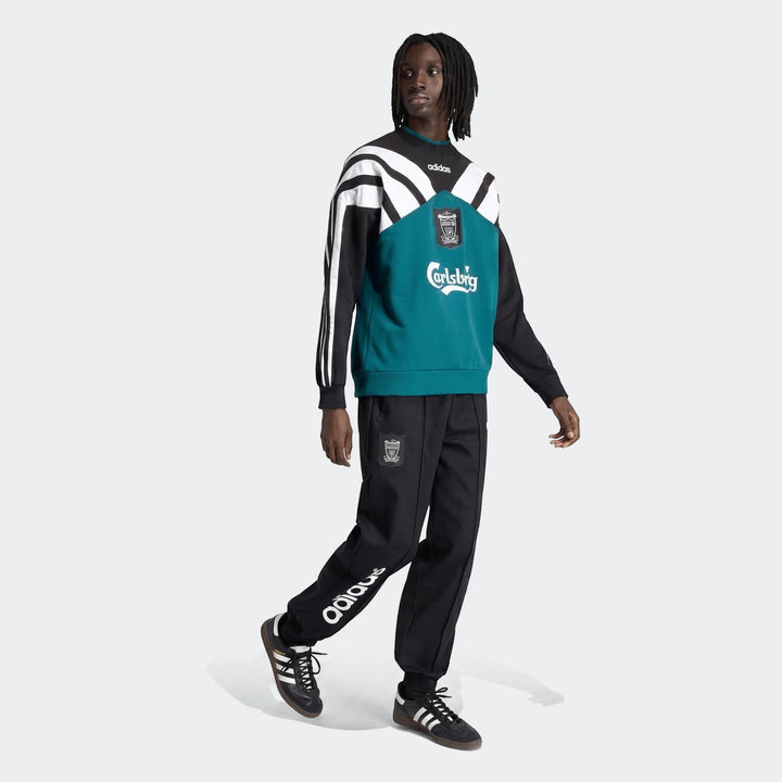 A male model wearing a black, white, and teal Liverpool Fc 1995 sweatshirt, black Adidas track pants, and black Adidas Samba