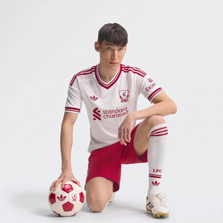 Male model in white and red Liverpool FC 26/27 Away Jersey Authentic, red shorts, and white socks, kneeling with a soccer