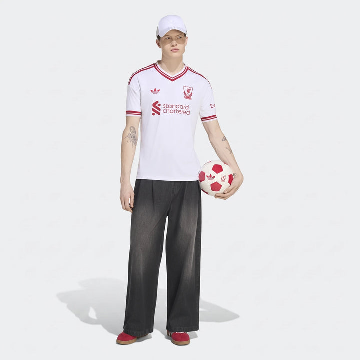 A male model wears the white Liverpool Fc 26/27 Away Jersey, black baggy jeans, and a white cap, holding a soccer ball.