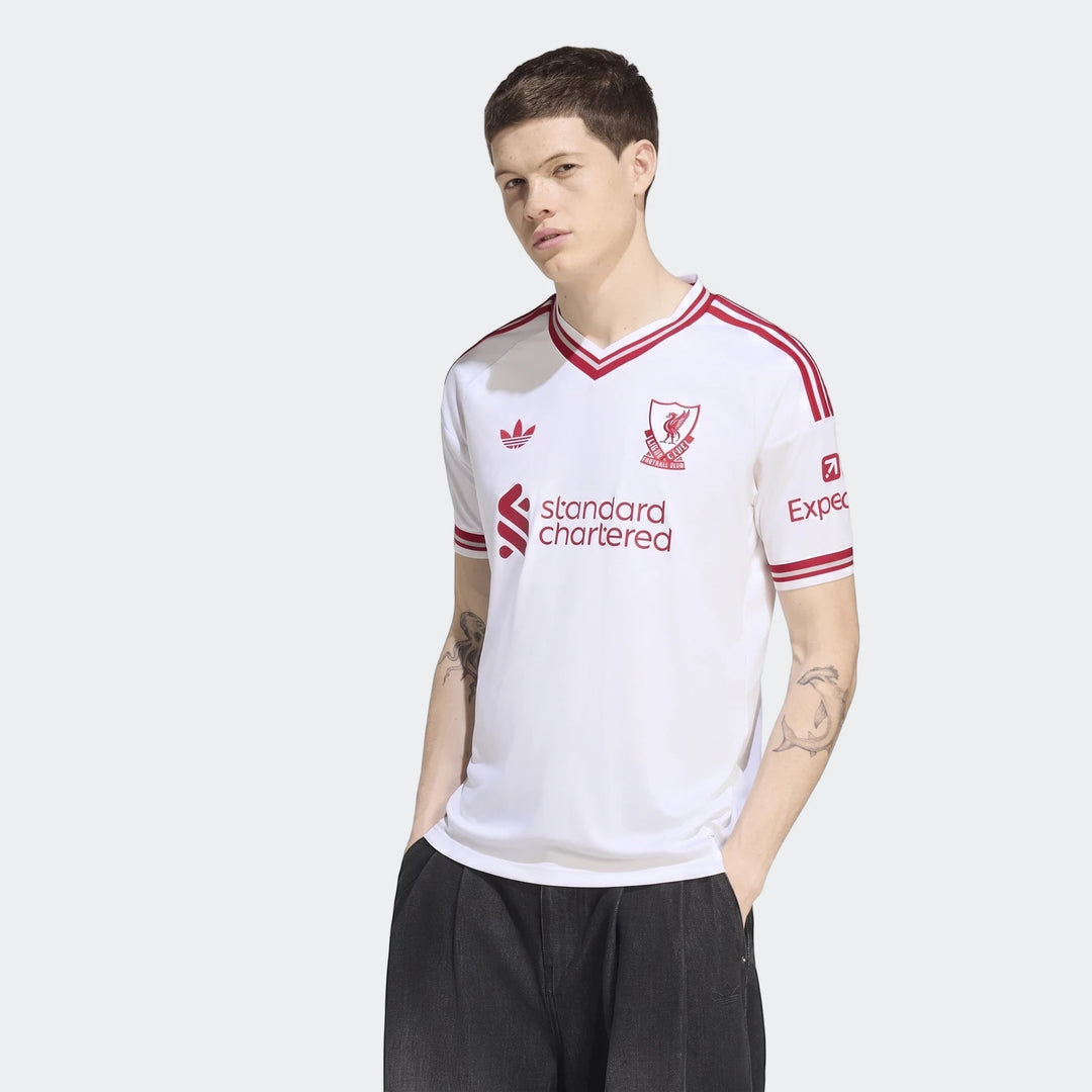 Male model wearing a white Liverpool FC 26/27 Away Jersey with red stripes and sponsor logos, hands in pockets.