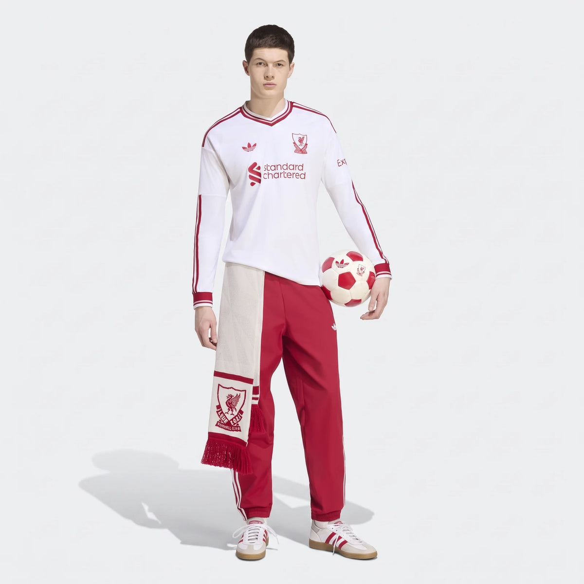 Male model wearing a white and red Liverpool FC 26/27 Away Longsleeve Jersey, red track pants, and holding a soccer ball and