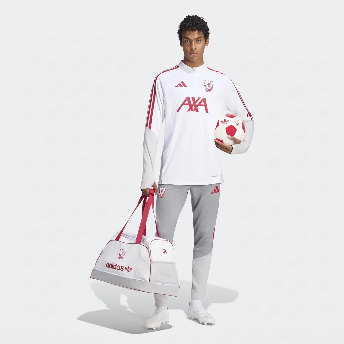 Male model wearing a white and red Liverpool FC 26/27 Cup Tiro Training Top and gray pants, holding a soccer ball and a