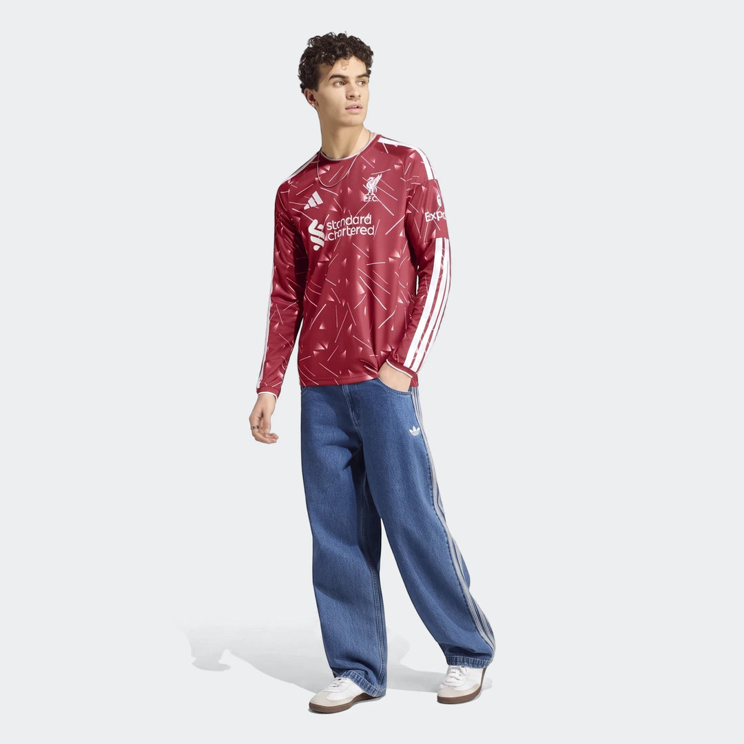 A male model wears a red Liverpool FC 26/27 home jersey with long sleeves, blue loose-fit jeans, and white sneakers, posing