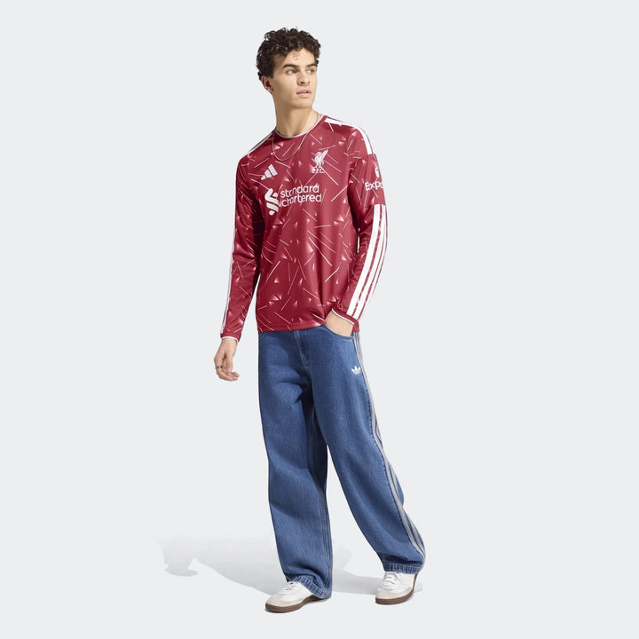 A male model wears a red Liverpool FC 26/27 home jersey with long sleeves, blue loose-fit jeans, and white sneakers, posing