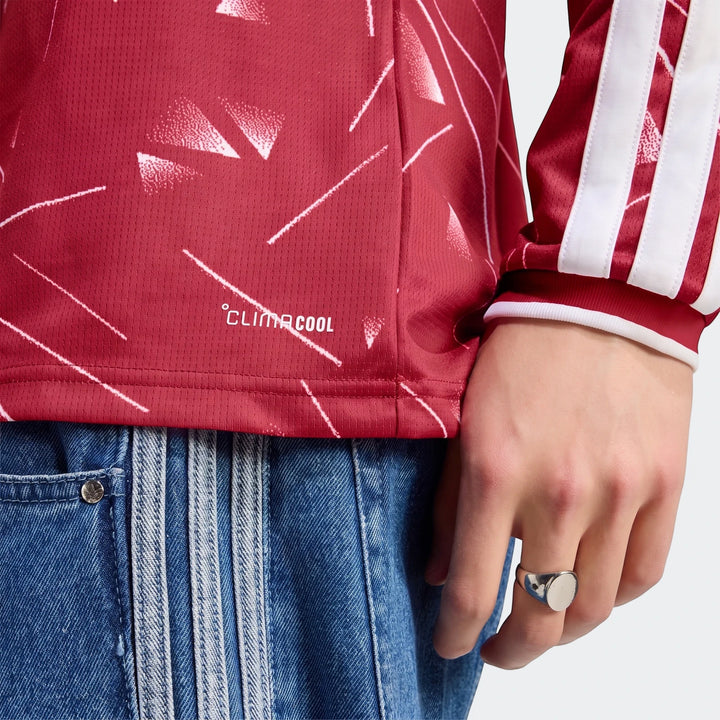 Close-up of a person wearing the red Liverpool FC 26/27 Home Long Sleeve Jersey with white geometric patterns and blue jeans.