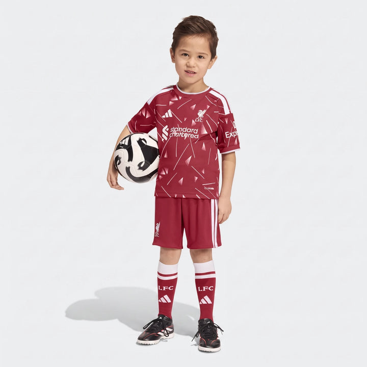 Young boy stands full body in a maroon Liverpool FC 26/27 Home Kids Set, featuring jersey, shorts, and socks, holding a