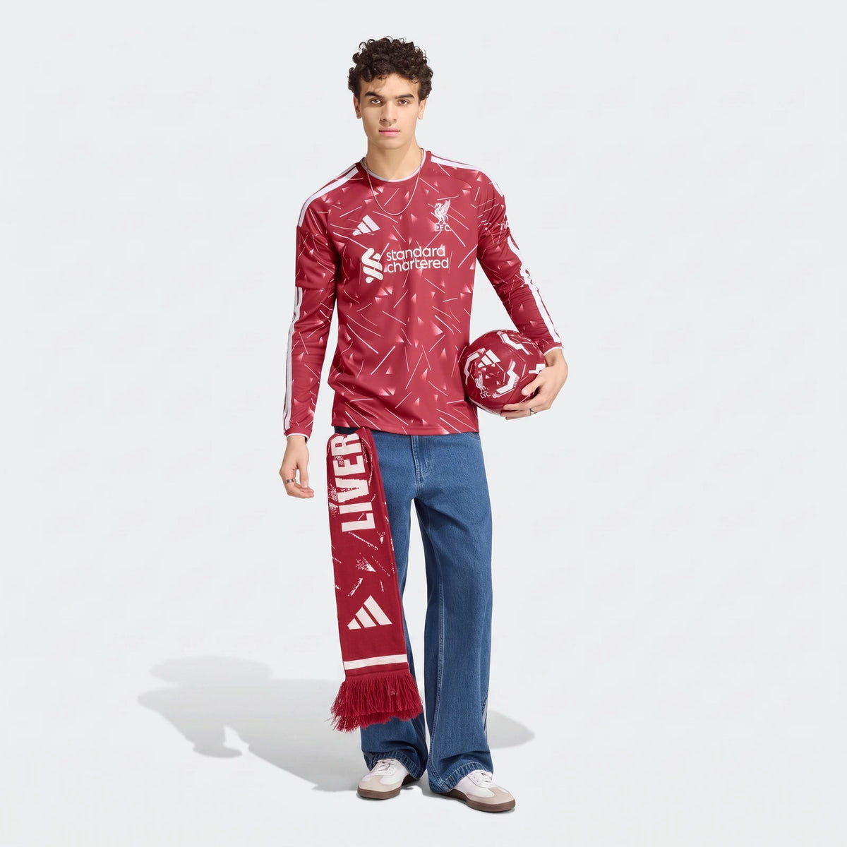 Male model wearing a red Liverpool FC 26/27 home long sleeve jersey with white patterns, holding a matching football and