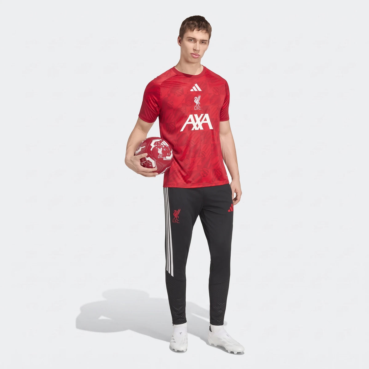 Male model wearing a red patterned Liverpool FC 26/27 Pre Match Jersey and black track pants, holding a red soccer ball.
